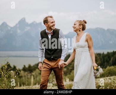 Gli sposi ridenti tengono le mani a piedi nelle montagne rocciose del Wyoming Foto Stock