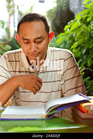 Smart del Sudest Asiatico giovane etnica adulto uomo davanti ad una scrivania, godere di studiare molti letteratura Foto Stock