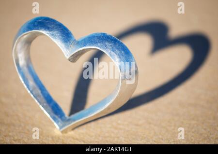 Cuore d'argento lucido in piedi sulla spiaggia che getta un'ombra su sabbia marrone Foto Stock