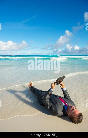 Uomo d'affari rilassato sdraiato in un vestito bagnato utilizzando il suo computer tablet sulla riva di una spiaggia tropicale Foto Stock