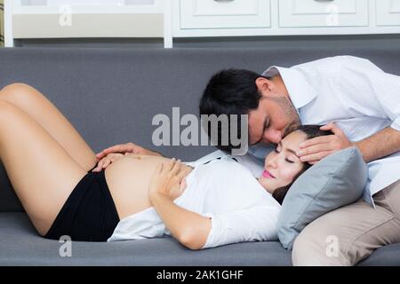 Il marito baciare la fronte della moglie incinta dalla pancia di mentre è seduto sul divano. Foto Stock