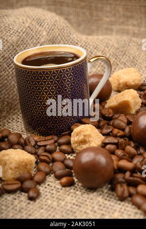 Una tazza di caffè, caffè in grani, pezzi di zucchero di canna e il cioccolato su uno sfondo di homespun ruvida stoffa. Close up Foto Stock