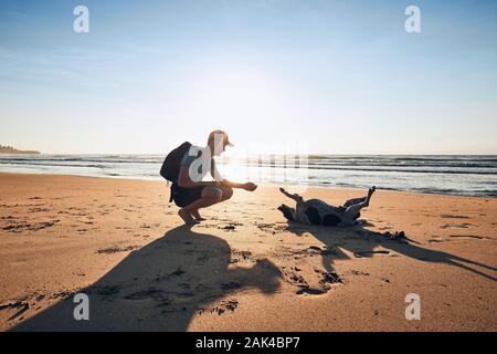 Giovane uomo giocando con allegro cane sulla spiaggia di sabbia contro il mare, Sri Lanka. Foto Stock