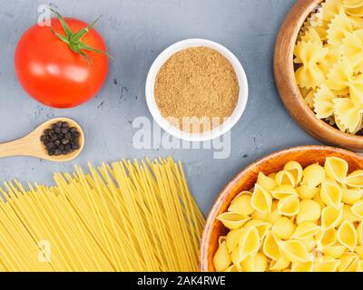 Diversi tipi di pasta in ciotole di legno, Masala, pomodoro, pepe nero su un cemento grigio Sfondo. Mangiare sano concetto Foto Stock