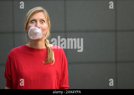 Donna bionda divertendosi con bubblegum o gomme da masticare soffiando una bolla di grandi dimensioni con occhi spalancati contro un muro grigio Foto Stock