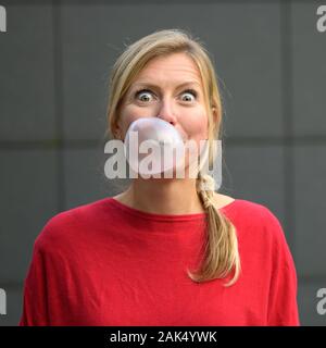 Donna bionda divertendosi con bubblegum o gomme da masticare soffiando una bolla di grandi dimensioni con occhi spalancati contro un muro grigio Foto Stock