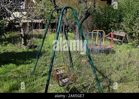 Vecchio altalene arrugginite merry-go-round e panca in legno in abbandonato incolto parco giochi per bambini. Foto Stock