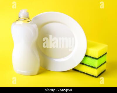 Bianco liquido per il lavaggio di stoviglie in una bottiglia di plastica, piastra e tre spugne di schiuma. Purezza e sostanze chimiche di uso domestico. Detergente da cucina su uno sfondo giallo. Foto Stock