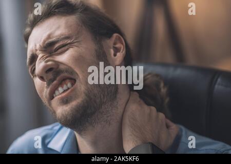 Dolorosa sensazione. Giovane uomo barbuto in maglietta blu che soffrono di dolori al collo Foto Stock