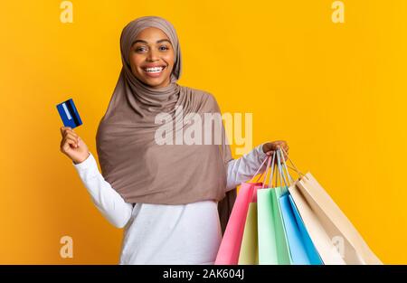 Bonus di rimborso concetto. Felice nero ragazza musulmana in hijab tenendo la carta di credito e colorato shopping bags su sfondo giallo, spazio libero Foto Stock