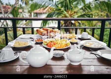 La colazione pronta da mangiare. Uova, frutta tropicale, toasts e tè pentola sul tavolo di legno contro il palm tree. Foto Stock