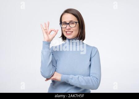 Caucasian donna matura con un ampio sorriso che indossano occhiali che mostra il simbolo ok con le dita. Il linguaggio del corpo okay significato super gesto per il negozio ottica, de Foto Stock