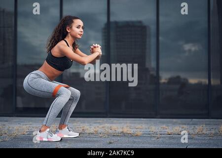 Atletica Giovane donna facendo squat esercizi con gomma sportive presso la città urbana ubicazione Foto Stock