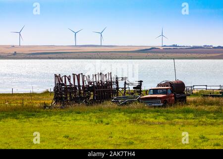 Un ampio angolo di visione di vecchie rusty farm equipment memorizzato in un campo sul lungomare. Vecchio camion e trattori gli allegati da un lago con le turbine eoliche in background Foto Stock