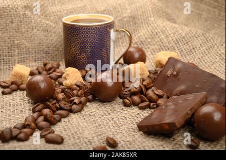 Una tazza di caffè, caffè in grani, pezzi di zucchero di canna e il cioccolato su uno sfondo di homespun ruvida stoffa. Close up Foto Stock