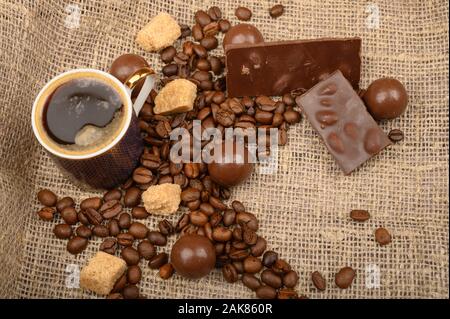 Una tazza di caffè, caffè in grani, pezzi di zucchero di canna e il cioccolato su uno sfondo di homespun ruvida stoffa. Close up Foto Stock