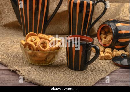 Piccole ciambelle, lo zucchero di canna, una tazza di tè e piatti in ceramica su uno sfondo di ruvida stoffa homespun close-up Foto Stock