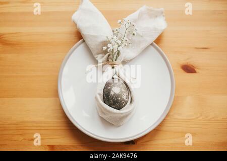 Tavola di pasqua decorazioni. Elegante brunch pasquale messa in tavola con uovo in Easter Bunny igienico. Moderno tinto naturale uovo di marmo sul tovagliolo con bunny orecchio Foto Stock