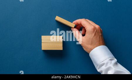 La mano di un uomo d affari con il posizionamento di un picchetto in legno sulla parte superiore della pila di essi in una immagine concettuale. Su sfondo blu con una copia dello spazio. Foto Stock