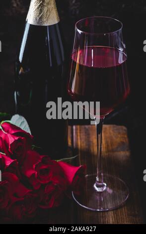 Impostazione scuro di un calice di vino e un mazzo di rose rosse con bottiglia in background su un tavolo di legno Foto Stock