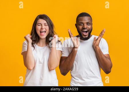 Interracial giovane gioia il successo. Overjoyed ragazza emotivamente stringendo i pugni con gli occhi chiusi mentre lei sconvolto il ragazzo nero alzando le mani Foto Stock