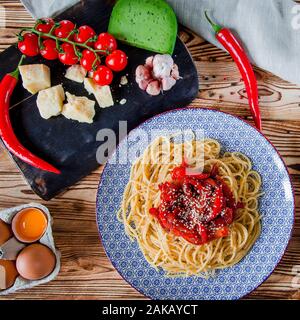Parmigiano pomodori ciliegia, peperoncino, Verde formaggio e aglio giacciono su un bordo scuro che sorge su di un tavolo di legno accanto a un bel piatto Foto Stock