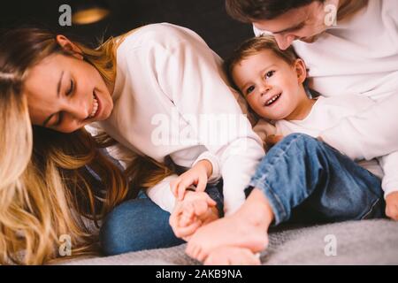 La famiglia felice è divertimento in camera da letto. Godendo di essere insieme. I genitori sono solletico il loro piccolo figlio mentre giaceva a letto. Famiglia allegra giocando Foto Stock