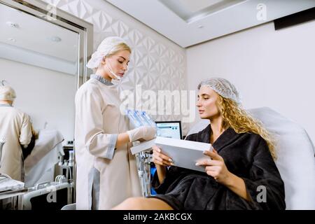 La cosmetologia cabinet, donna seduta client sul lettino. Estetista sorge e mostra il trattamento cosmetici iniezioni. Cosmetologo e discutere del paziente Foto Stock