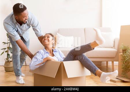 Spostando un nuovo appartamento. Gioiosa African American uomo Moglie di equitazione in una scatola di cartone interna dopo il trasferimento. Messa a fuoco selettiva Foto Stock