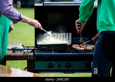 Un uomo la cottura di salsicce su un gas aperto barbeque, vi è anche un vassoio di cipolle in attesa di essere servito Foto Stock