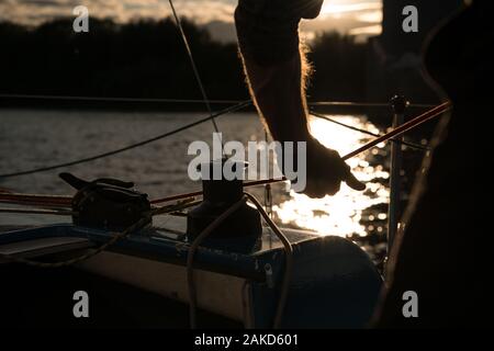 Primo piano di una mano che tiene una corda a prua di una barca vela su un fiume sul tramonto. Foto Stock