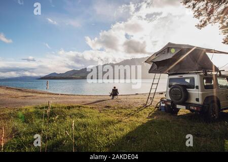 Una donna siede lungo un lago accanto ad un SUV con una tenda in Nuova Zelanda Foto Stock