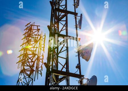 Una vista astratta di due stazioni base cellulari torri, reticolo in acciaio design con le comunicazioni elettroniche ingranaggio su una giornata soleggiata con colorate lens flare Foto Stock