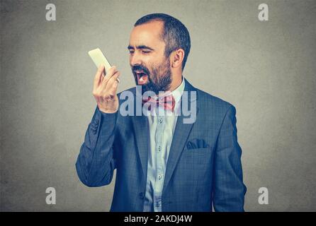 Ritratto arrabbiato uomo barbuto che indossa elegante abito rosso con il filtro bow tie, urlando, gridando mentre si parla al telefono, isolato, grigio Sfondo. Negativo Foto Stock