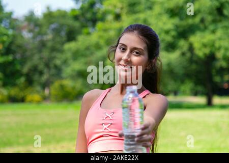 Sorridente giovane ragazza adolescente in tuta sportiva bere e dare una bottiglia d'acqua ad un amico per una buona vita sana dopo l'esecuzione di esercizio nel parco su estate weeke Foto Stock