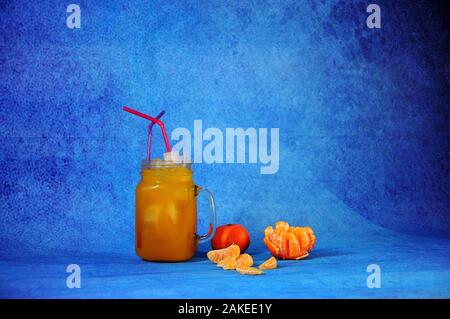 Una tazza di succo di mandarino con ghiaccio e cannucce accanto a pelati e tutta la frutta e fette di mandarino su uno sfondo blu. Close-up. Foto Stock