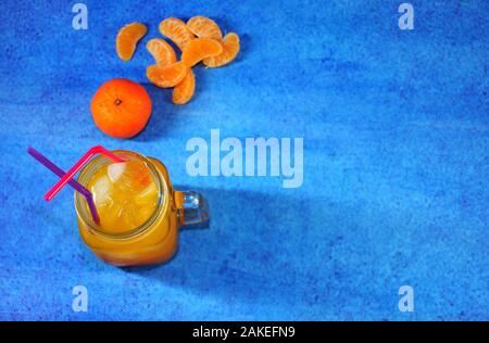 Una tazza di succo di mandarino con ghiaccio e cannucce accanto a pelati e tutta la frutta e fette di mandarino su uno sfondo blu. Close-up. Foto Stock