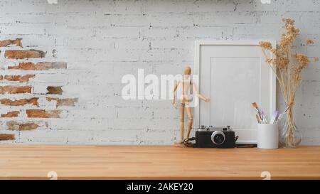 Ritagliato colpo di designer contemporanei workplace con fotocamera, legno figura, il telaio e le spazzole sul tavolo di legno con un muro di mattoni sfondo Foto Stock