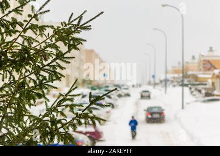 L'inverno è di nevicare, rami di abete rosso contro lo sfondo di una strada innevata, scura di auto e un pedone in blu sono mobili lungo il parcheggio, Foto Stock