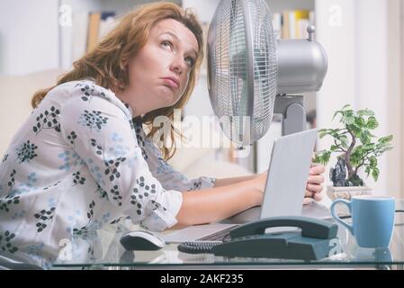 La donna soffre di calore mentre si lavora in ufficio e si tenta di raffreddarsi dalla ventola Foto Stock