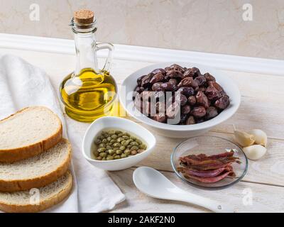 Ingredienti per la tapenade: indurite o secche le olive in un recipiente bianco, acciughe, capperi, aglio, olio di oliva e il pane su una tavola di legno. Cucina mediterranea Foto Stock