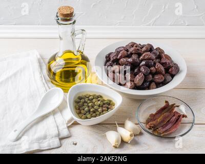 Ingredienti per la tapenade: indurite o secche le olive in un recipiente bianco, filetto di acciuga, capperi, aglio e olio di oliva su un tavolo di legno. Cucina mediterranea. Foto Stock