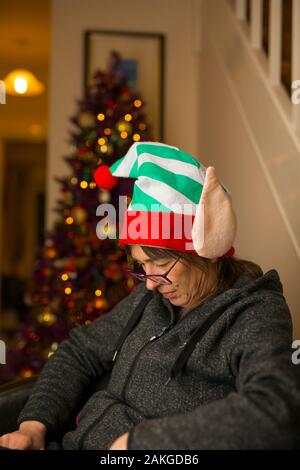 Divertente close up di mezza età britannico donna caucasica isolata, addormentarsi nella sedia la pancia gonfia dopo aver mangiato il pranzo di Natale che indossa silly elf hat. Foto Stock