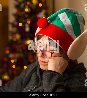 Divertente close up di mezza età britannico donna caucasica isolata, addormentarsi nella sedia la pancia gonfia dopo aver mangiato il pranzo di Natale che indossa silly elf hat. Foto Stock