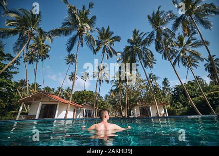 Relax in piscina nel mezzo di palme di cocco. Giovane uomo appoggiato in acqua contro i bungalow del complesso turistico. Foto Stock