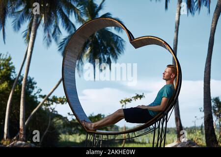 Relax in un paradiso tropicale. Giovane uomo seduto nel cuore di legno contro le palme e il paesaggio tropicale. Foto Stock