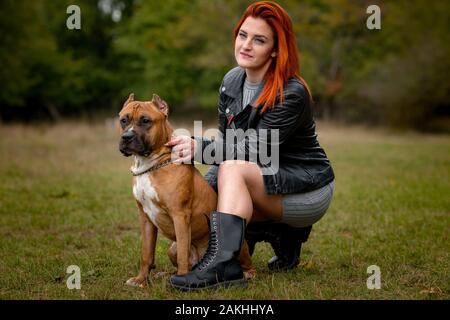 Bella donna e il suo cane nel parco di caduta. Bello cane American Staffordshire Terrier in piedi vicino a donna in piedi in erba verde. Foto Stock