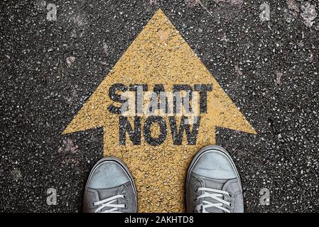 Elevato angolo di visione della persona in piedi sulla freccia su asfalto, su strada con il testo inizia ora, il concetto di motivazione Foto Stock