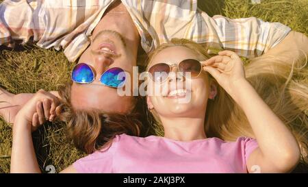 Felice coppia giovane sdraiato sul prato, tenendo le mani, godendo di fine settimana nel giorno di estate Foto Stock