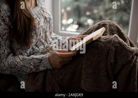 Attraente ragazza è la lettura di libro seduti sul davanzale della casa. Ampia finestra, piante verdi. Foto Stock